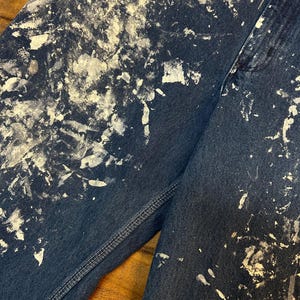 May include: A pair of blue denim jeans with white paint splatters. The jeans have a straight leg and a classic five-pocket design.