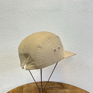 May include: A tan-colored, five-panel cap with a short brim. The cap has two small ventilation eyelets on each side and a stitched seam detail. The hat is displayed on a metal stand against a neutral background.