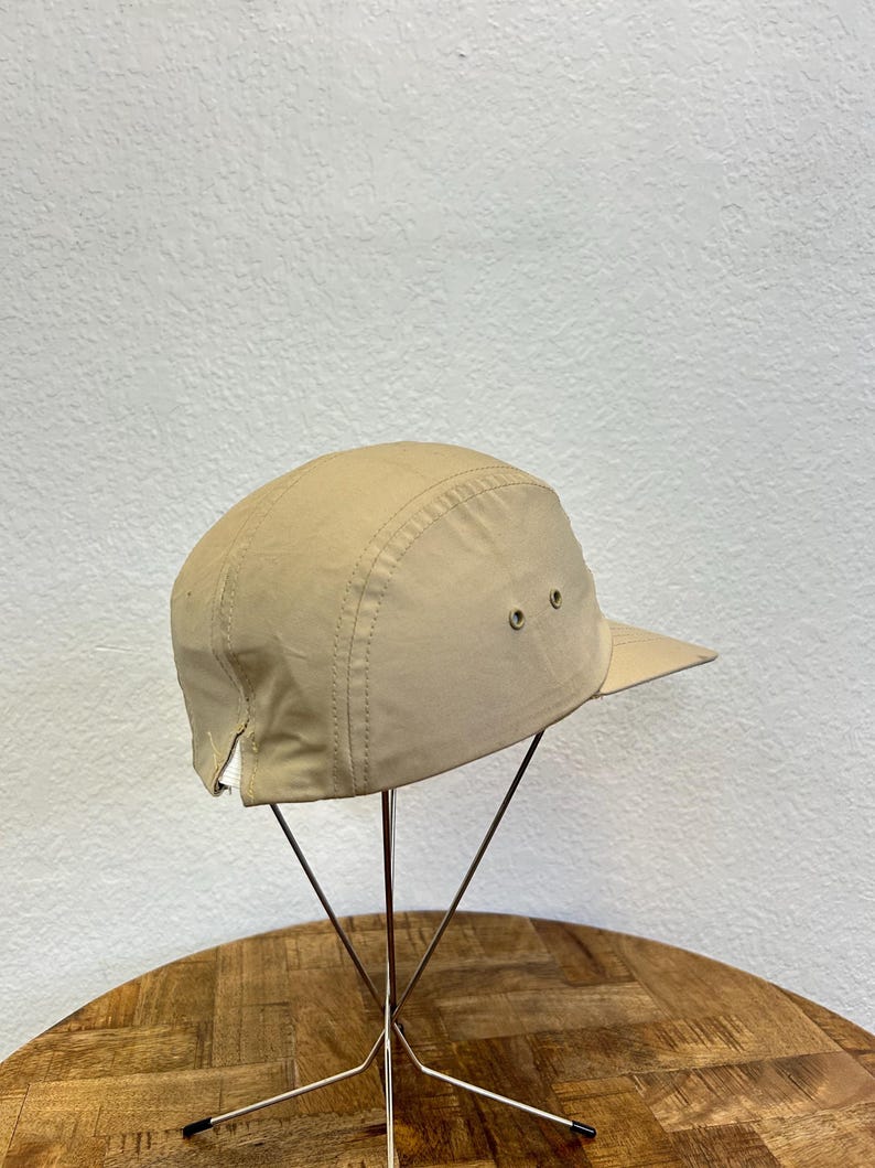 May include: A tan-colored, five-panel cap with a short brim. The cap has two small ventilation eyelets on each side and a stitched seam detail. The hat is displayed on a metal stand against a neutral background.