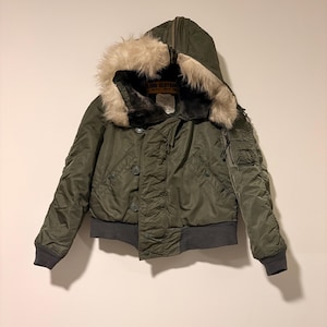 Könnte beinhalten: Olivgrüne Bomberjacke mit fellgefütterter Kapuze. Die Jacke hat einen gerippten Saum und Bündchen, eine Knopfleiste und ein Etikett mit der Aufschrift "GLOBE CLOTHING". Die Jacke ist auf Wärme und Stil ausgelegt.