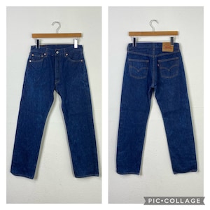 May include: A pair of blue denim jeans hanging on a wooden hanger. The jeans have a classic straight leg style and are made of a sturdy, durable fabric. The jeans have a red leather patch on the back pocket with the Levi's logo.