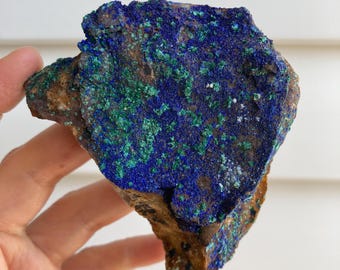 May include: A rough-hewn rock formation featuring a vibrant display of blue and green mineral deposits. The stone has a brown base with patches of deep blue and emerald green hues, creating a striking contrast. The rock is held in a hand.