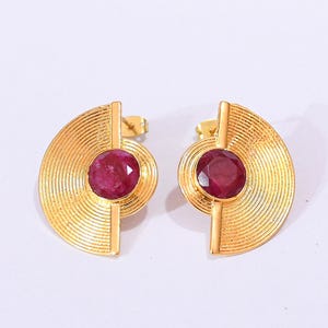 Pode incluir: Um par de brincos dourados com um design de leque semicircular e linhas concêntricas finas. Cada brinco é centrado por uma pedra preciosa redonda, facetada e de cor vermelho-escura, fixada por uma barra metálica vertical no centro.