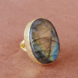 May include: A gold ring with a large oval-shaped labradorite gemstone. The gemstone has a blue, green, and brown iridescent sheen.