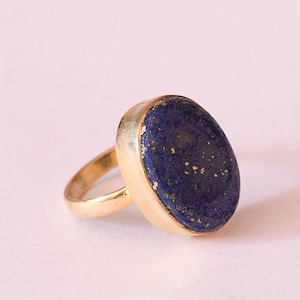 Puede incluir: Un anillo de oro con una gran piedra ovalada de lapislázuli. La piedra azul oscuro tiene motas doradas, engastada en una banda de oro pulido. El anillo es una pieza llamativa.