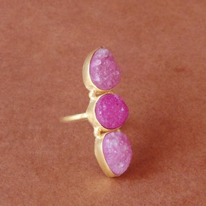 May include: A gold ring with three bright pink druzy stones. The stones are set vertically, displaying a textured, crystalline surface. The ring's band and settings have a polished finish. The ring is a statement piece.