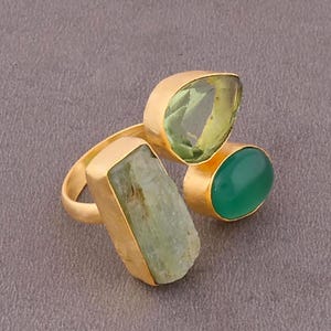 Peut inclure: Une bague dorée ornée de trois pierres précieuses : une pierre en forme de goutte d'eau vert clair, une pierre verte foncée arrondie et une pierre rectangulaire vert clair. La bague a un design ouvert unique.