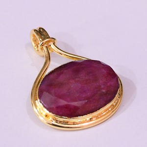 Op de afbeelding: Een gouden hanger met een grote, facetgeslepen, ovale robijn. De robijn heeft een diepe, rijke rode kleur en is gezet in een gouden frame met een decoratieve balein voor een ketting. De hanger is voor sieraden.