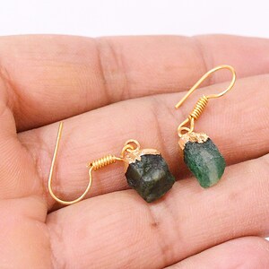 Puede incluir: Un par de pendientes dorados con piedras preciosas en bruto de color verde oscuro. Los pendientes tienen un diseño de gancho simple con un acento enrollado cerca de la piedra. Las piedras preciosas tienen una textura natural.