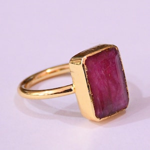 May include: A gold ring with a rectangular, deep red gemstone. The gemstone is set in a gold bezel, and the band is a simple, polished gold. The ring is shown against a soft, light background.