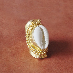 Könnte beinhalten: Ein goldfarbener Ring mit einem weißen Muschelschalen-Mittelstück. Der Ring hat ein breites Band mit dekorativen Perlen- und Seil-Details. Die Muschelschale ist oval geformt und hat eine glatte, weiße Oberfläche. Der Ring ist vor einem rötlich-braunen Hintergrund platziert.