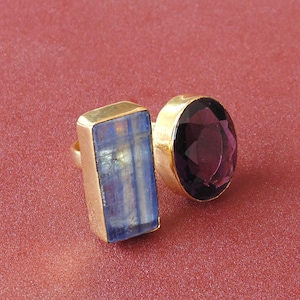 May include: Two gold rings with large gemstones. One ring features a rectangular blue gemstone, and the other has an oval, deep purple gemstone. The rings are set against a reddish-pink background.