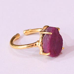 Könnte beinhalten: Ein goldener Ring mit einem großen, rohen, violetten Edelstein, der in einer einfachen goldenen Fassung sitzt.