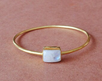 May include: A gold-coloured bangle bracelet featuring a rectangular, iridescent, white gemstone at the centre. The bracelet is set against a solid, muted pink background. The gemstone has a subtle blue and white shimmer.