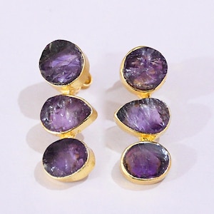 Puede incluir: Un par de pendientes dorados con tres piedras de amatista moradas en bruto. Las piedras están engastadas en un bisel dorado y conectadas por eslabones dorados. Los pendientes tienen un diseño colgante y cierre de poste.