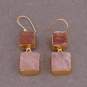 Puede incluir: Pendientes de tono dorado con dos piedras cuadradas. La piedra superior es de color marrón rojizo y la piedra inferior es de color rosa claro.