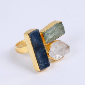 May include: A gold-toned ring featuring three raw gemstones: a rectangular blue stone, a rectangular green stone, and a clear, irregular-shaped stone. The ring has a simple band and a gold setting.