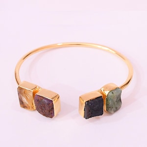 May include: A gold-toned cuff bracelet featuring four raw, colorful gemstones. The gemstones are set in gold-colored rectangular bezels. The bracelet has a simple, open design, with the gemstones as the focal point.