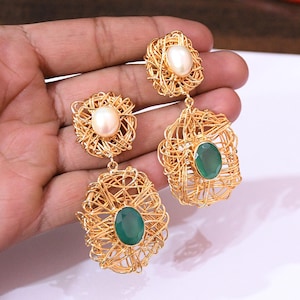Può includere: Orecchini in filo dorato con un design intrecciato unico. Ogni orecchino presenta una perla o una gemma ovale verde. Gli orecchini sono tenuti da una mano, mostrando l'intricata lavorazione artigianale e il contrasto di colori.
