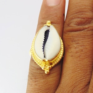 Könnte beinhalten: Ein goldfarbener Ring mit einer weißen und violetten Kaurimuschel. Die Muschel ist in einen dekorativen Goldrahmen mit einem seilartigen Rand gefasst. Der Ring wird an einem Finger gezeigt.
