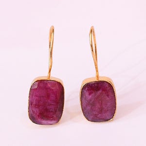 May include: A pair of gold-toned earrings featuring large, rectangular, faceted, deep magenta gemstones. The gemstones are set in gold bezels and suspended from simple gold ear wires. The earrings are displayed against a soft, off-white background.
