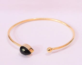 May include: A gold-toned open cuff bracelet featuring a smooth, polished metal band. One end of the bracelet is finished with a small, round gold-toned bead, while the other end is accented with a single, faceted, teardrop-shaped black stone.