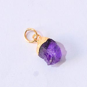 Puede incluir: Un colgante de amatista en bruto con una tapa y un asa doradas. El cristal púrpura tiene una textura rugosa y natural. El asa dorada está unida a la parte superior de la amatista, lista para ser ensartada en una cadena.