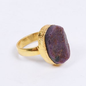 Może przedstawiać: Złoty pierścionek z dużym, surowym, fioletowo-czerwonym kamieniem szlachetnym. Kamień ma nieregularny kształt i jest osadzony w dekoracyjnym, teksturowanym obramowaniu. Obręcz pierścionka jest gładka, kontrastując z ozdobną oprawą.