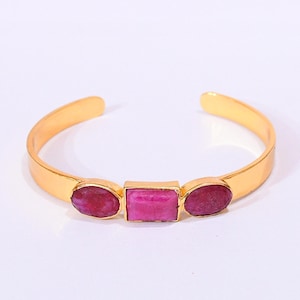 May include: A gold-toned open cuff bracelet featuring three bezel-set stones. The centre stone is a rectangular deep pink ruby, flanked by two oval-shaped deep pink rubies. The metal band is smooth, polished, and has a wide, open-ended design.
