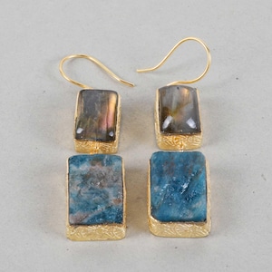 May include: A pair of gold-coloured drop earrings. Each earring features a rectangular gemstone, with one pair in blue and the other in a mix of grey, purple, and gold. The gemstones are set in ornate gold-coloured frames.