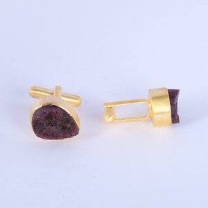 May include: A pair of gold-toned cufflinks with a natural, dark red-purple stone inlay. The cufflinks have a classic design with a rectangular and teardrop shape, showcasing the unique texture of the stone. The background is plain white.