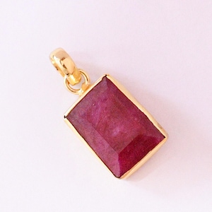 May include: A rectangular pendant featuring a deep magenta-colored gemstone set in a gold-toned metal frame. The gemstone has a faceted surface and is attached to a small, circular gold-toned bail at the top for hanging on a chain or cord.