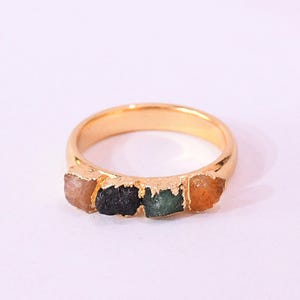 Könnte beinhalten: Ein goldfarbener Ring mit vier rohen, farbigen Edelsteinen. Die Edelsteine sind in den Farbtönen Orange, Grün und Schwarz gehalten. Der Ring ist vor einem weißen Hintergrund platziert.