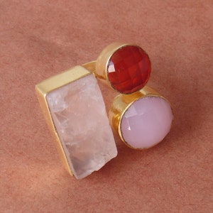 May include: A gold-toned ring featuring three gemstones: a rectangular, light pink stone, a round, faceted red stone, and a round, faceted light pink stone. The ring has a simple, elegant design.