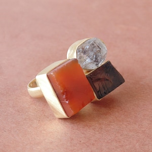 Op de afbeelding: Een gouden ring met drie geometrische edelstenen: een heldere, gefacetteerde steen, een rood-oranje vierkant en een donkerbruin vierkant. De ring heeft een geborstelde gouden afwerking, op een roze achtergrond.