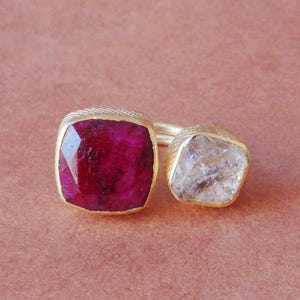 May include: A gold ring featuring two gemstones. One is a faceted, square-cut ruby, and the other is a clear, irregularly shaped stone. The ring has a textured band and a gold setting, set against a pink background.