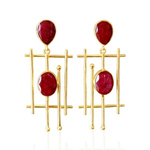 Op de afbeelding: Een paar goudkleurige oorbellen met een geometrisch ontwerp. Elke oorbel heeft een druppelvormige, dieprode edelsteen aan de bovenkant en een ovale edelsteen in het midden. De oorbellen hebben een lineaire, kooi-achtige structuur.