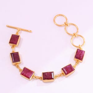 May include: A gold-tone bracelet featuring five rectangular, deep red faceted stones set in metal frames. The bracelet includes a toggle clasp on one end and a series of three circular links on the other end to allow for adjustable sizing.