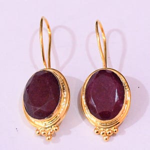 Op de afbeelding: Een paar goudkleurige oorbellen met ovale, facetgeslepen, dieprode edelstenen. De edelstenen zijn gezet in gouden bezels met een decoratieve cluster van kleine gouden kralen aan de onderkant. De oorbellen hebben een eenvoudig, gebogen haakontwerp.