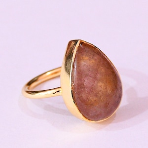 May include: A gold-colored ring featuring a teardrop-shaped, reddish-brown gemstone. The gemstone is set in a gold bezel, and the ring band is also gold. The ring is displayed against a white background.