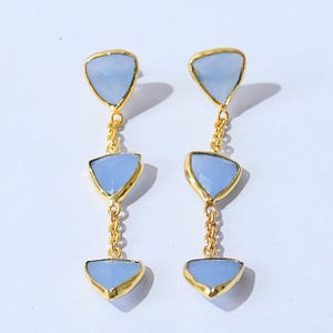 Peut inclure: Une paire de boucles d'oreilles pendantes avec trois pierres triangulaires bleu clair serties dans du métal doré. Les pierres sont reliées par de fines chaînes dorées. Les boucles d'oreilles ont un design simple et élégant.