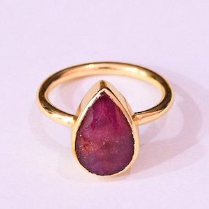 Może przedstawiać: Złoty pierścionek z gruszkowym, ciemnoczerwonym kamieniem szlachetnym. Obręcz i oprawa pierścionka są złote, co kontrastuje z żywym kamieniem szlachetnym. Pierścionek to biżuteria.