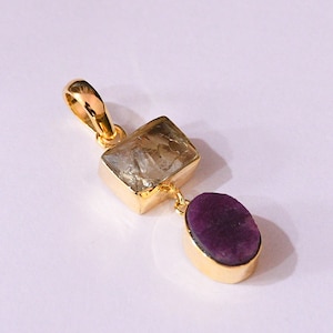 Könnte beinhalten: Ein goldfarbener Anhänger mit zwei Edelsteinen. Der obere Stein ist ein rechteckiger, hellfarbiger Kristall, und der untere Stein ist ein tiefvioletter Oval. Der Anhänger hat eine Schlaufe für eine Kette.