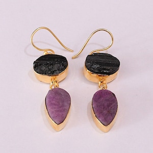 May include: A pair of gold-colored earrings. Each earring features a black, rough-textured stone at the top, with a teardrop-shaped, purple stone hanging below. The stones are set in gold-colored metal.