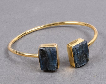 May include: Gold-tone metal cuff bracelet featuring two rectangular blue gemstone accents. The bracelet has an open design, with the gemstones set in gold-coloured bezels. The gemstones have a natural, textured appearance.