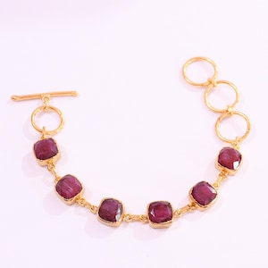 May include: A gold-toned bracelet featuring five square-cut, deep magenta-colored gemstones set in metal bezels. The bracelet includes a toggle clasp closure on one end and three circular metal links on the other end, connected by small gold-toned chains.