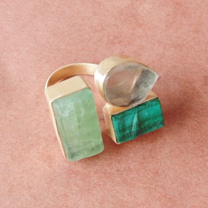 May include: A gold ring with three gemstone settings. It features a rectangular, light green stone, a clear, teardrop-shaped stone, and a rectangular, green malachite stone. The ring is set against a pink background.