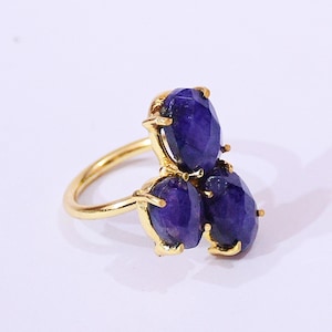 May include: A gold ring with three oval, deep blue gemstones. The gemstones are arranged in a cluster, with a polished gold band and settings. The ring is presented against a white backdrop. The gemstones are approximately 1 cm in length.