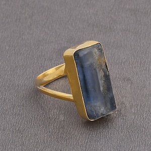 Pode incluir: Um anel dourado com uma pedra preciosa azul retangular. A pedra tem uma aparência natural e texturizada com tons de azul e cinza. O anel tem um design de haste simples e dividida.