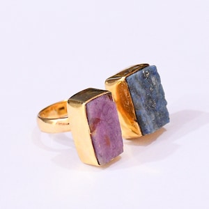 May include: Two gold-plated rings with rectangular gemstone settings. One ring features a purple gemstone, the other a blue gemstone. The rings are displayed on a white background, showcasing the jewelry's design and color.